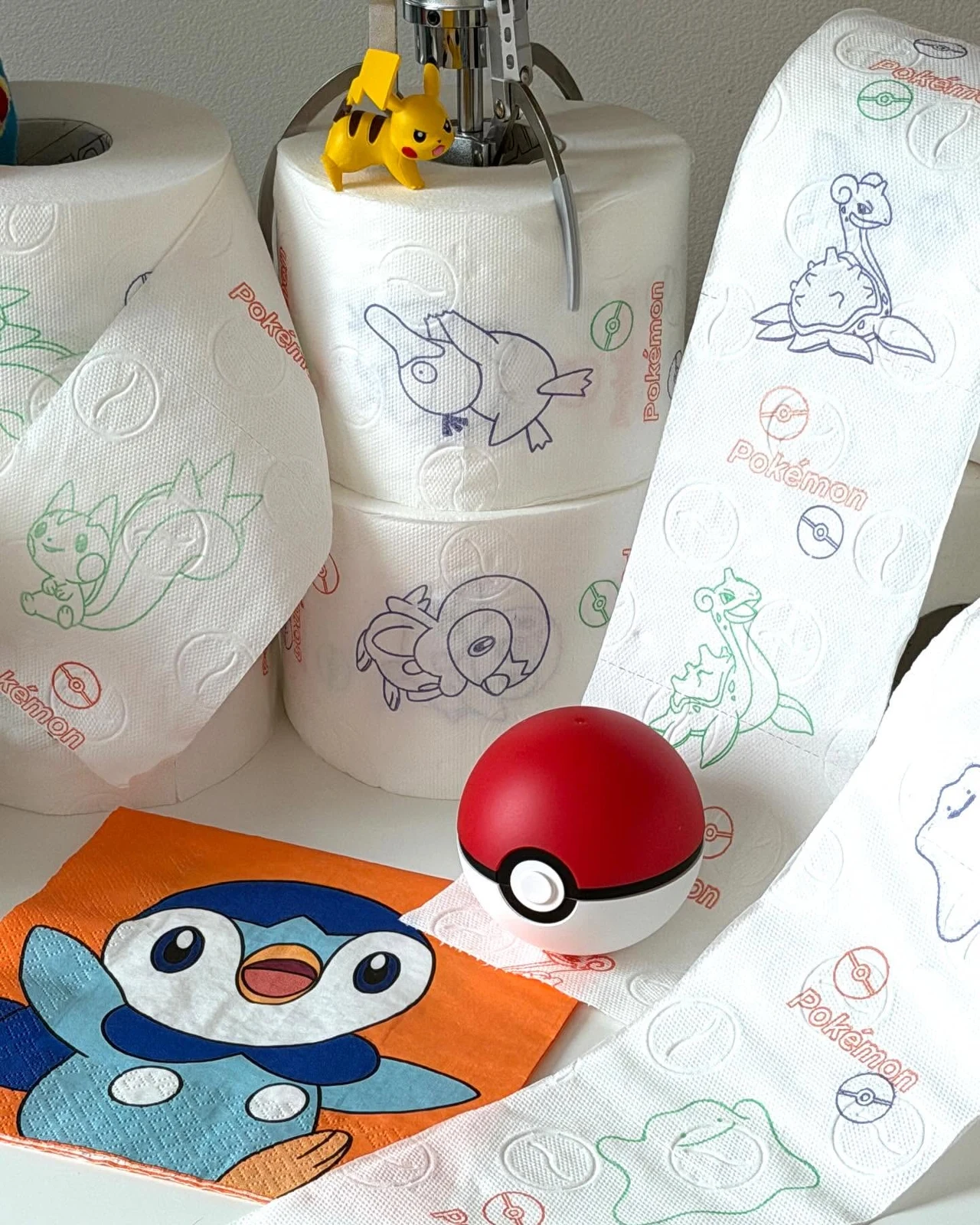 Biały papier toaletowy z kolorowymi nadrukami postaci Pokémon, pomarańczowa serwetka z wizerunkiem Piplupa, figurka Pikachu i czerwono-biała kula Pokéball.