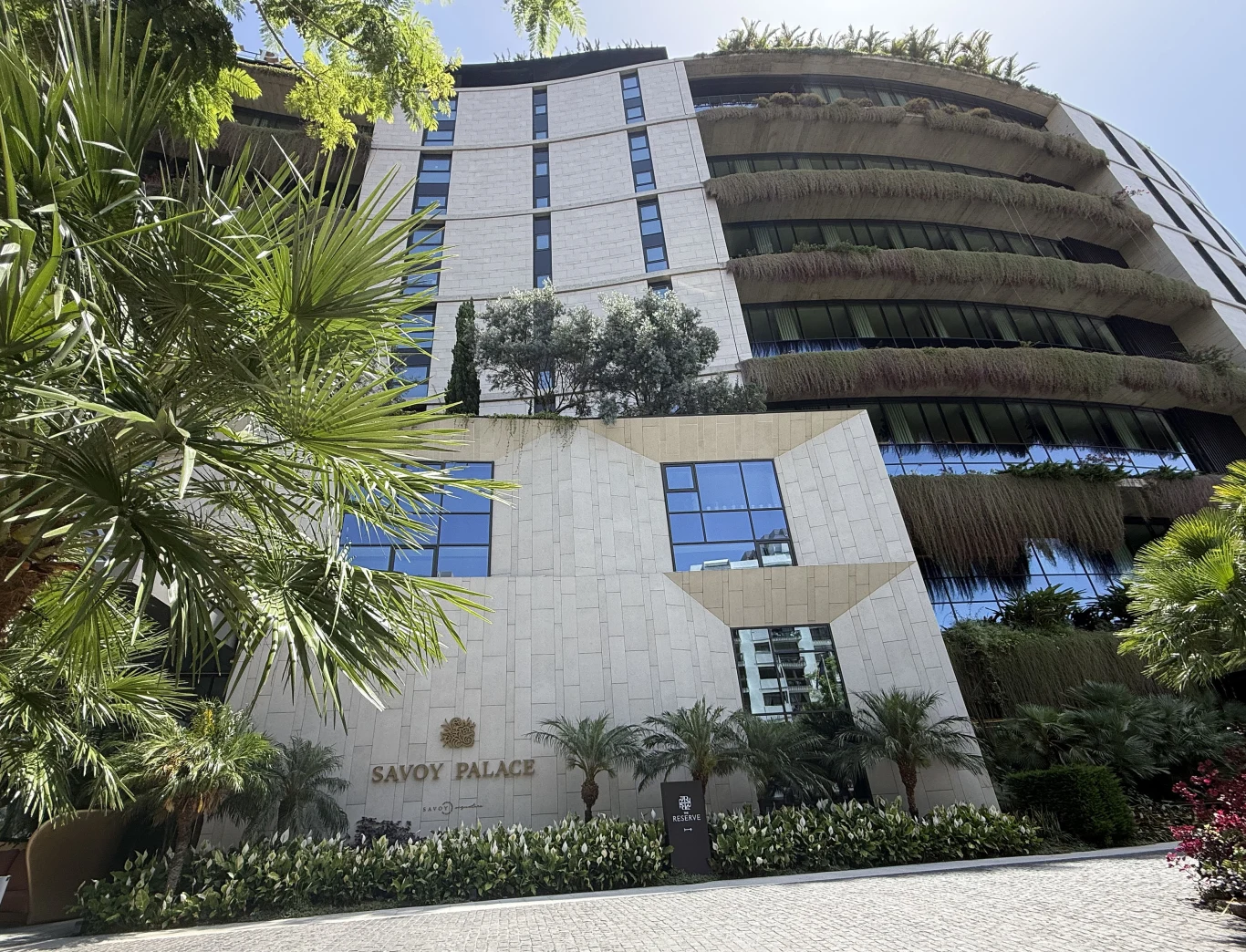 Hotel Savoy Palace w Funchal, stolicy należącej do Portugalii wyspy Madera Fasada nowoczesnego budynku hotelowego z geometrycznymi kształtami, dużymi przeszklonymi oknami i zielenią na balkonach, otoczona palmami i egzotycznymi roślinami.