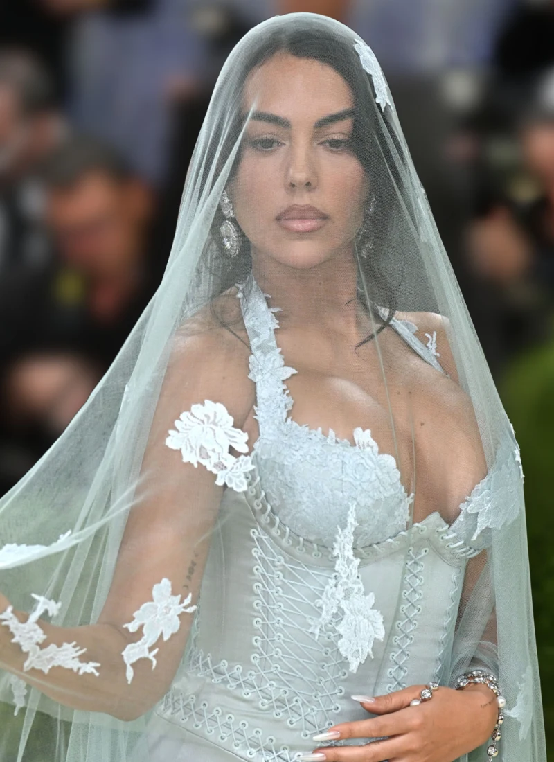 Georgina Rodriguez Kobieta w eleganckiej, jasnej sukni z gorsetem i kwiatowymi aplikacjami, okryta transparentnym welonem, patrzy wprost przed siebie.