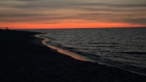 Linia brzegowa plaży o zmierzchu z ciemnym piaskiem i łagodnymi falami, nad którymi rozciąga się pomarańczowo-różowe niebo zachodzącego słońca.