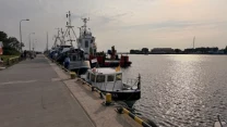 Rząd zacumowanych łodzi i kutrów przy nabrzeżu portowym, tafla wody odbijająca światło oraz widoczna część infrastruktury portowej w oddali.