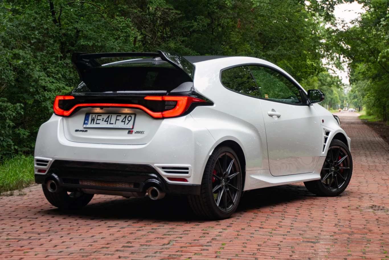 Biała sportowa Toyota GR Yaris z dużym tylnym spojlerem, czarnymi felgami i podwójnym wydechem stoi na czerwonej brukowanej drodze otoczonej zielonymi drzewami.