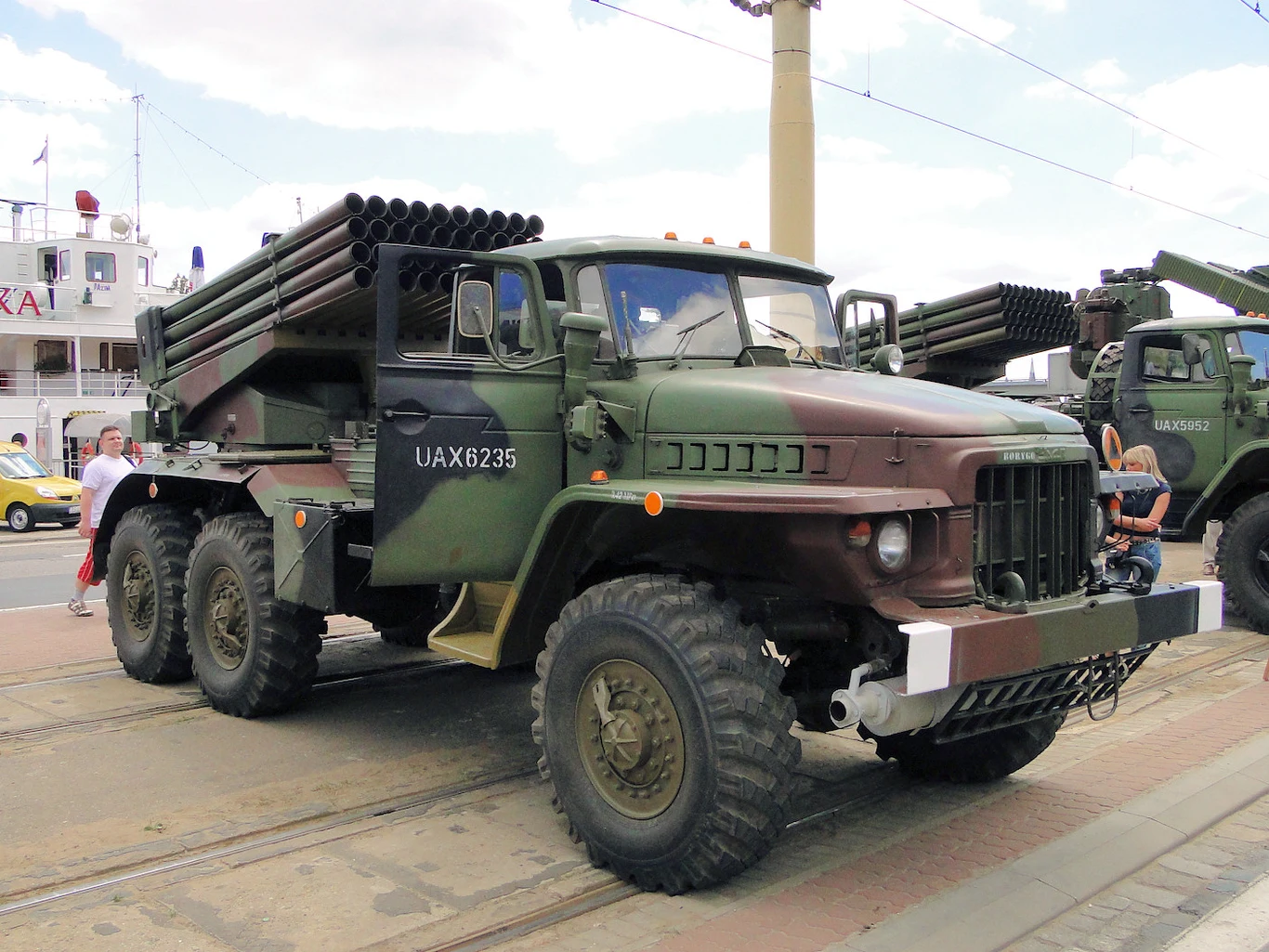 BM-21 Grad z zasobów Wojska Polskiego. Wojskowa ciężarówka w kamuflażu z zamontowaną wieloprowadnicową wyrzutnią rakiet, ustawiona na ulicy w otoczeniu ludzi i innych pojazdów militarnych.