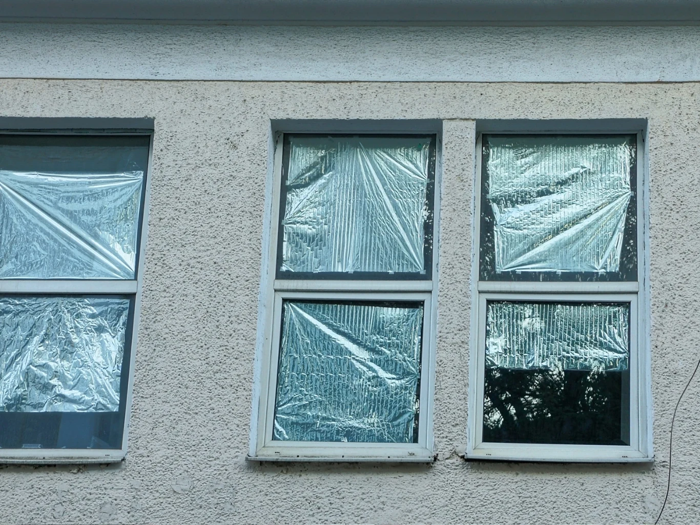 Folię zazwyczaj montuje się od środka, ale lepiej gdy jest po zewnętrznej stronie szyby, by jej nie uszkodzić Cztery okna w ścianie budynku zabezpieczone folią, co sugeruje prace remontowe lub ochronę przed wpływami zewnętrznymi.
