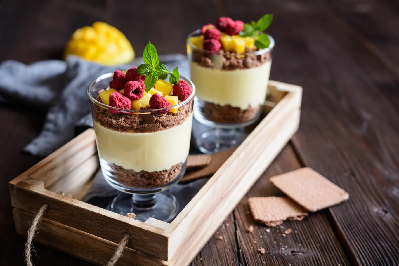Deserki w kubeczkach to smakowita propozycja na przysmak bez pieczenia Dwie szklanki deseru warstwowego z kremem, ciasteczkami, malinami, mango i miętą na drewnianej tacy z krakersami.