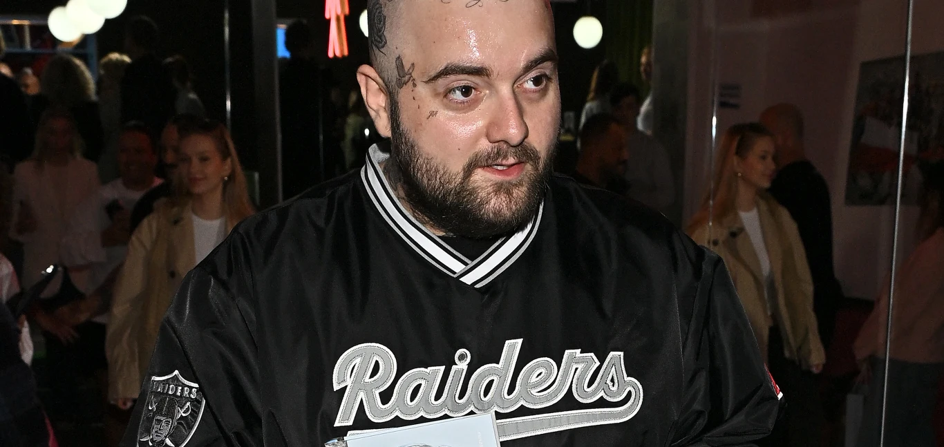 Bedoes Bedoes z tatuażami na twarzy w czarnej kurtce „Raiders” trzyma jasny przedmiot wśród ludzi w pomieszczeniu.