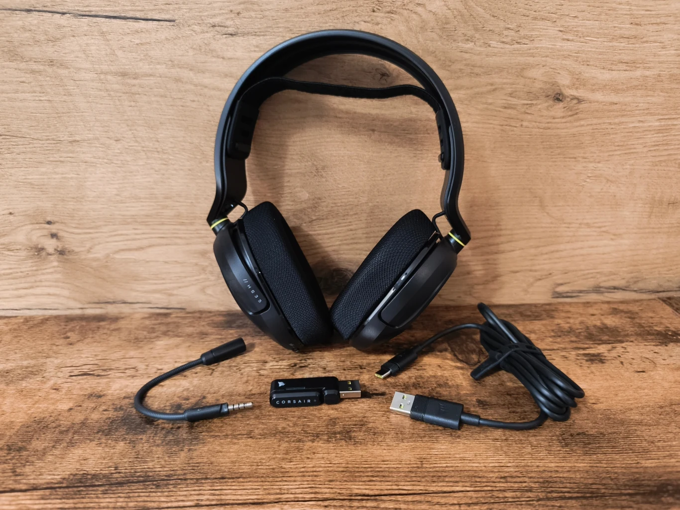Corsair HS35 v3 Wireless Czarne słuchawki nauszne z dołączonym odłączanym mikrofonem, przewodem USB oraz adapterem, umieszczone na drewnianej powierzchni.