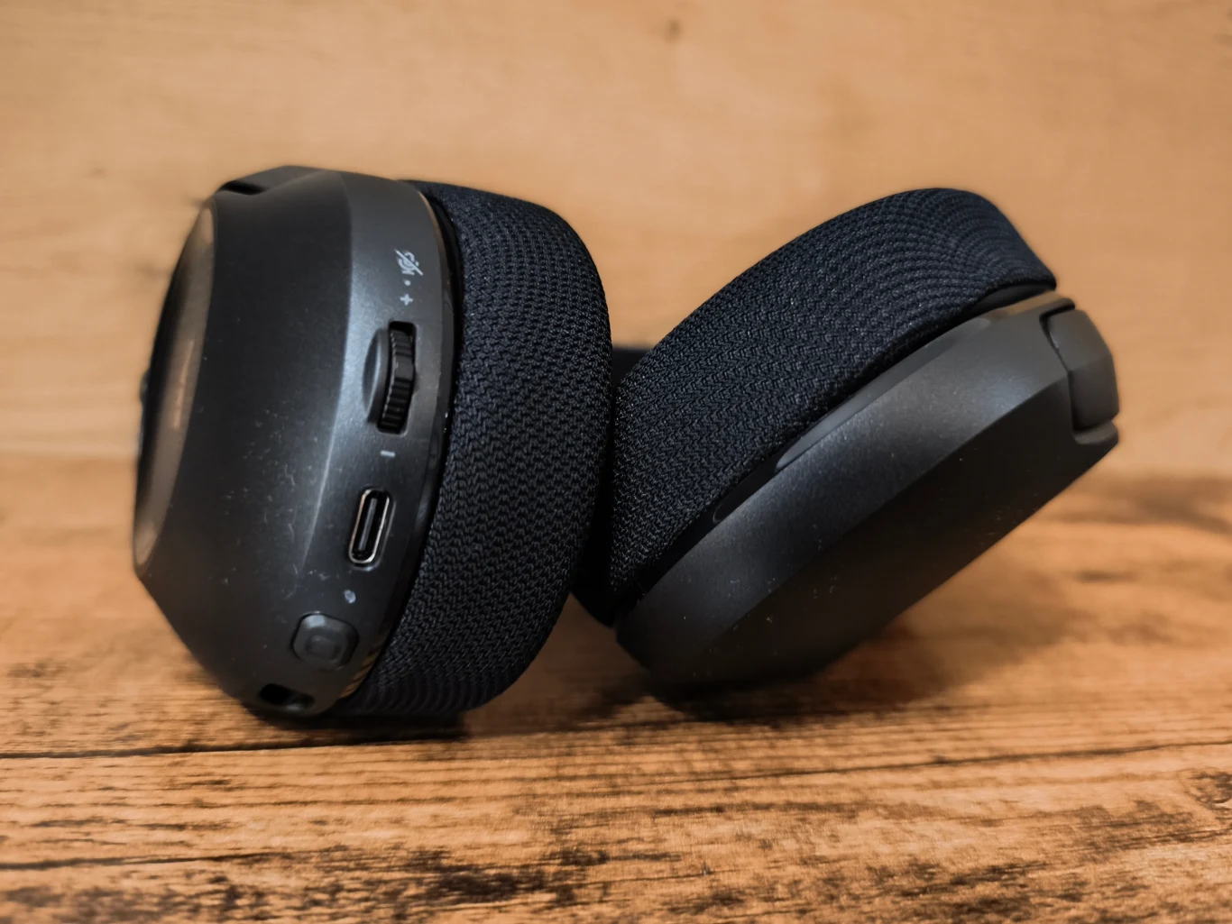 Corsair HS35 v3 Wireless Czarne nauszniki słuchawek z tekstylną poduszką oraz widocznym złączem USB-C i przyciskami sterującymi na jednej z muszli, ustawione na drewnianej powierzchni.