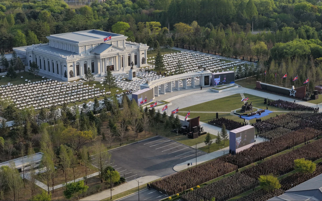 Muzeum Pamięci Czynów Bojowych Zagranicznych Operacji Wojskowych Monumentalny budynek w klasycznym stylu z dużą ilością kolumn, otoczony rzędami ustawionych krzeseł oraz zwartym zgromadzeniem ludzi ubranych na ciemno, skupionych wokół sceny z flagami i mównicą, całość otoczona bujną zielenią drzew.