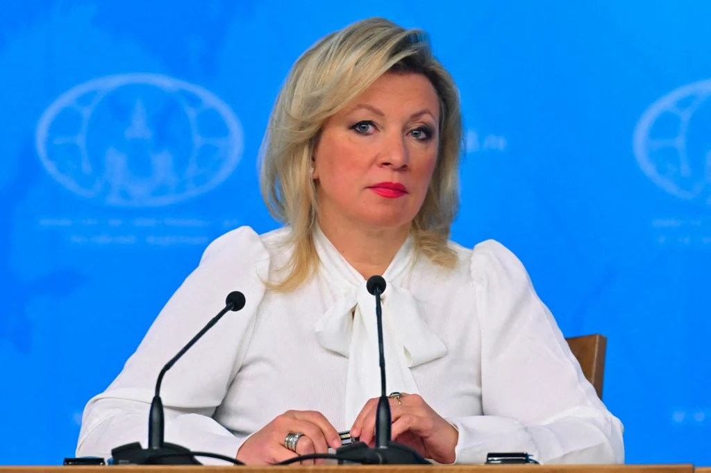 Maria Zacharowa Maria Zacharowa w białej bluzce siedzi przy stole z dwoma mikrofonami, na tle niebieskiej ściany z symbolem.