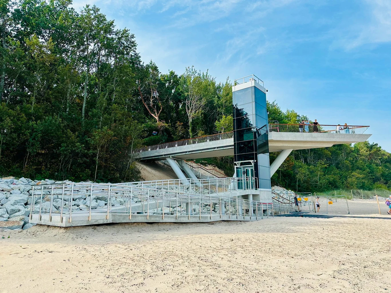 W Pobierowie ukończono realizację nietypowego zejścia na plażę / fot. Gmina Rewal Nowoczesne zejście na plażę w Pobierowie z tarasem, windą i podestem, otoczone drzewami.