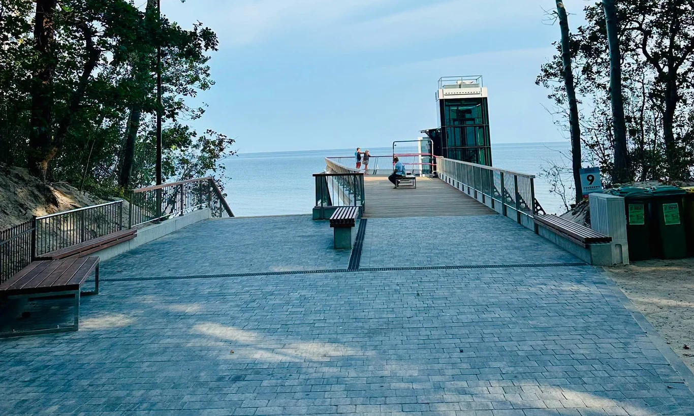 W Pobierowie powstało wyjątkowe zejście na plażę / fot. Gmina Rewal Szeroki deptak prowadzący do tarasu widokowego z windą, skierowanego ku morzu, otoczony drzewami i wyposażony w ławki oraz kosze na śmieci po prawej stronie.