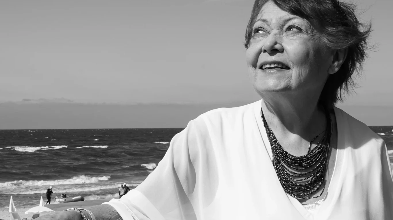 Iga Cembrzyńska (1939-2026) Iga Cembrzyńska w jasnej bluzce i naszyjniku nad morzem, patrzy w górę i uśmiecha się.