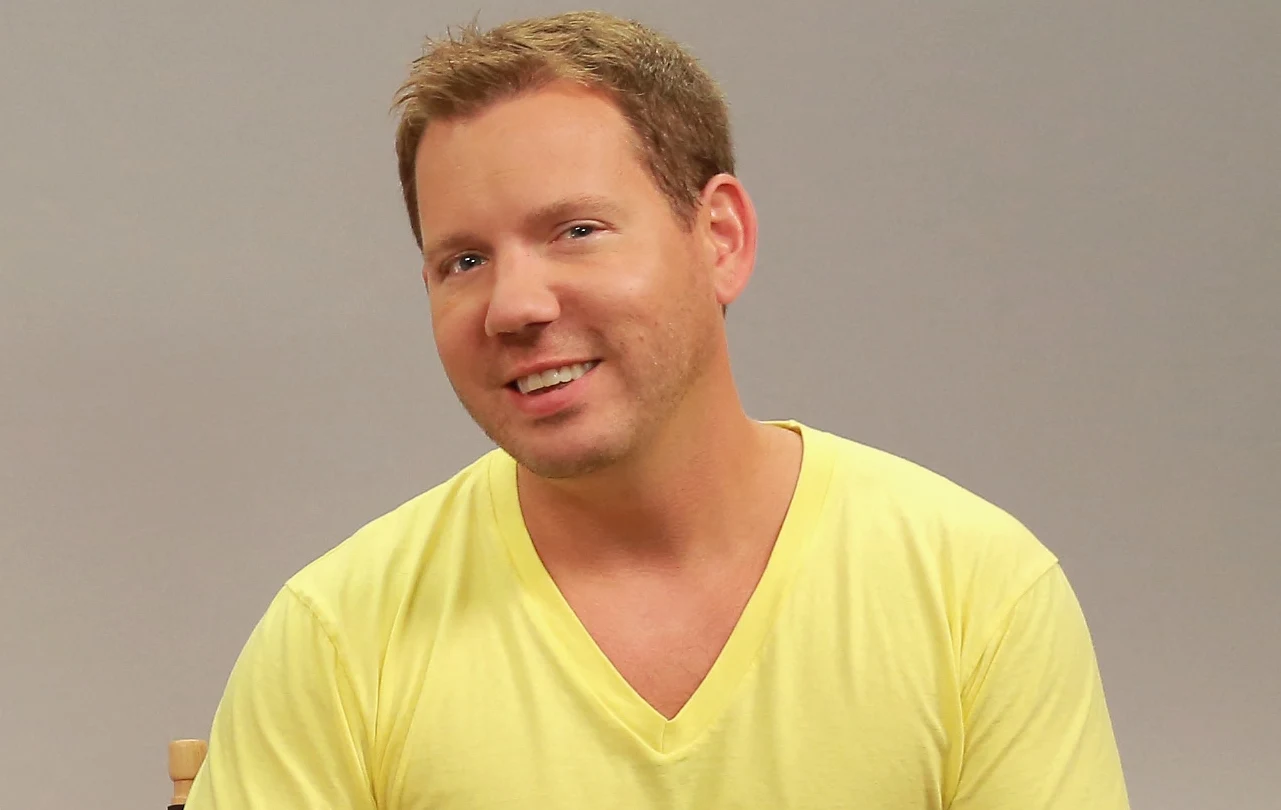 Cliff Bleszinski podczas panelu dyskusyjnego na Comic-Con 2014 Uśmiechnięty mężczyzna w żółtej koszulce z dekoltem w serek na neutralnym, szarym tle.