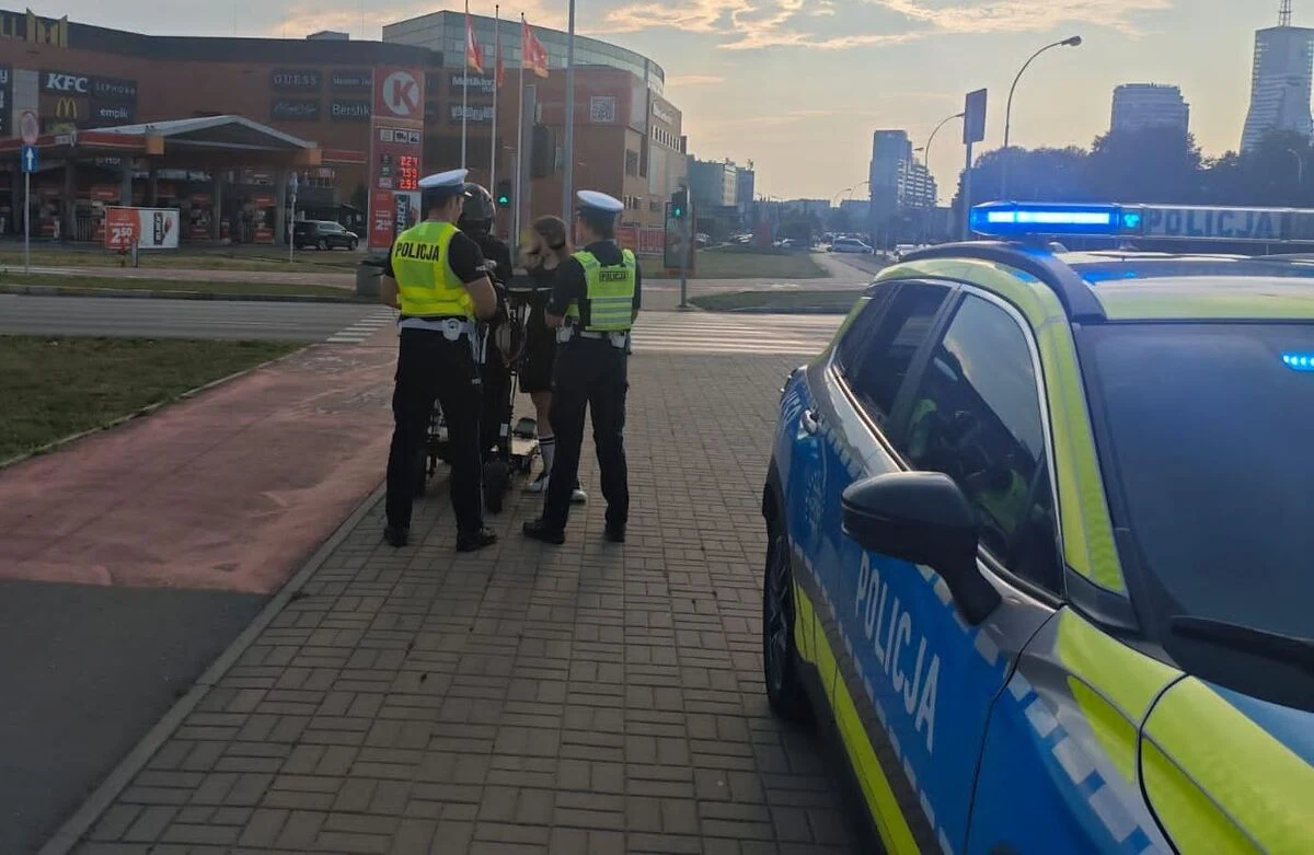 Policja udaremniła nielegalny rajd na hulajnogach w Rzeszowie Dwóch policjantów w kamizelkach odblaskowych przy hulajnodze elektrycznej, obok radiowóz policyjny w mieście.