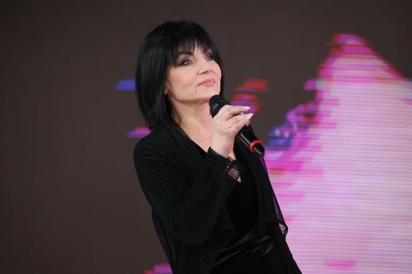 Shazza to legenda disco polo Shazza, kobieta o ciemnych włosach w czarnym stroju, trzyma mikrofon na tle kolorowej, rozmytej grafiki.