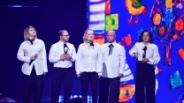 Pięć osób ubranych w białe koszule i czarne spodnie stoi na scenie, trzymając mikrofony, z kolorową, abstrakcyjną grafiką w tle.
