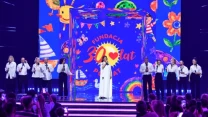 Grupa osób ubranych na biało wykonuje utwór na scenie, w tle kolorowa grafika z napisem „Fundacja 30 lat Pomagamy” oraz motywami dziecięcymi i rysunkami słońca, kwiatów i żaglówek, publiczność obserwuje wydarzenie i robi zdjęcia.
