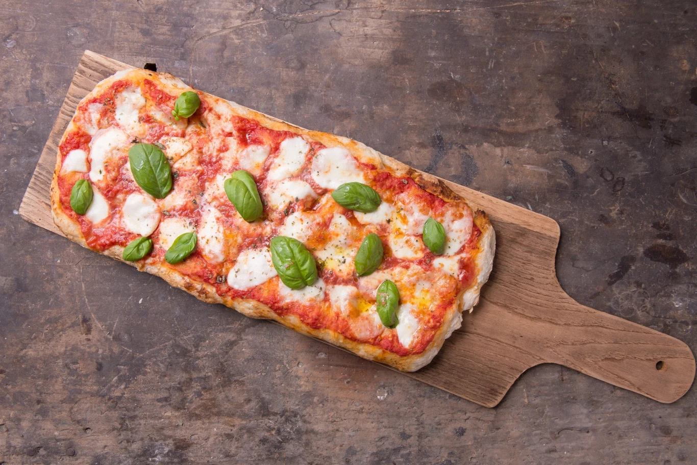 Pinsa Prostokątna pizza z sosem pomidorowym, roztopioną mozzarellą i świeżymi listkami bazylii, podana na drewnianej desce.