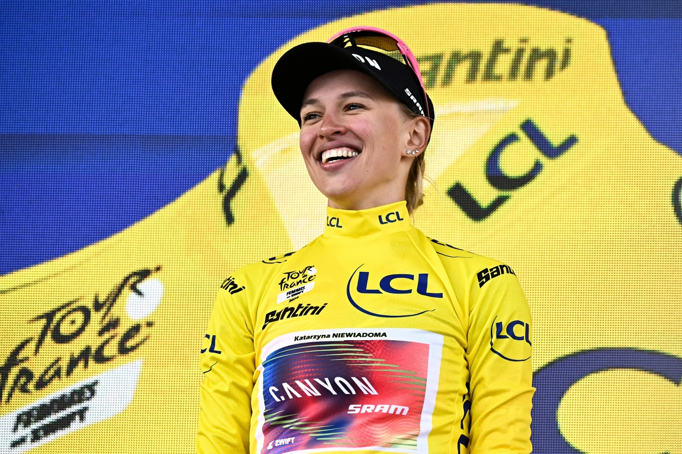 Katarzyna Niewiadoma-Phinney Kobieta ubrana w żółtą koszulkę lidera z logo LCL i Canyon Sram, uśmiechnięta, stoi na tle żółtej grafiki Tour de France Femmes.