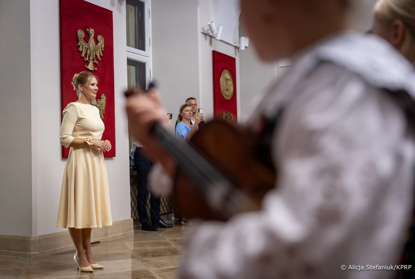 Elegancko ubrana kobieta stoi naprzeciwko dziecka grającego na skrzypcach w jasnym wnętrzu, ozdobionym biało-czerwonymi bannerami z orłem i innymi godłami.