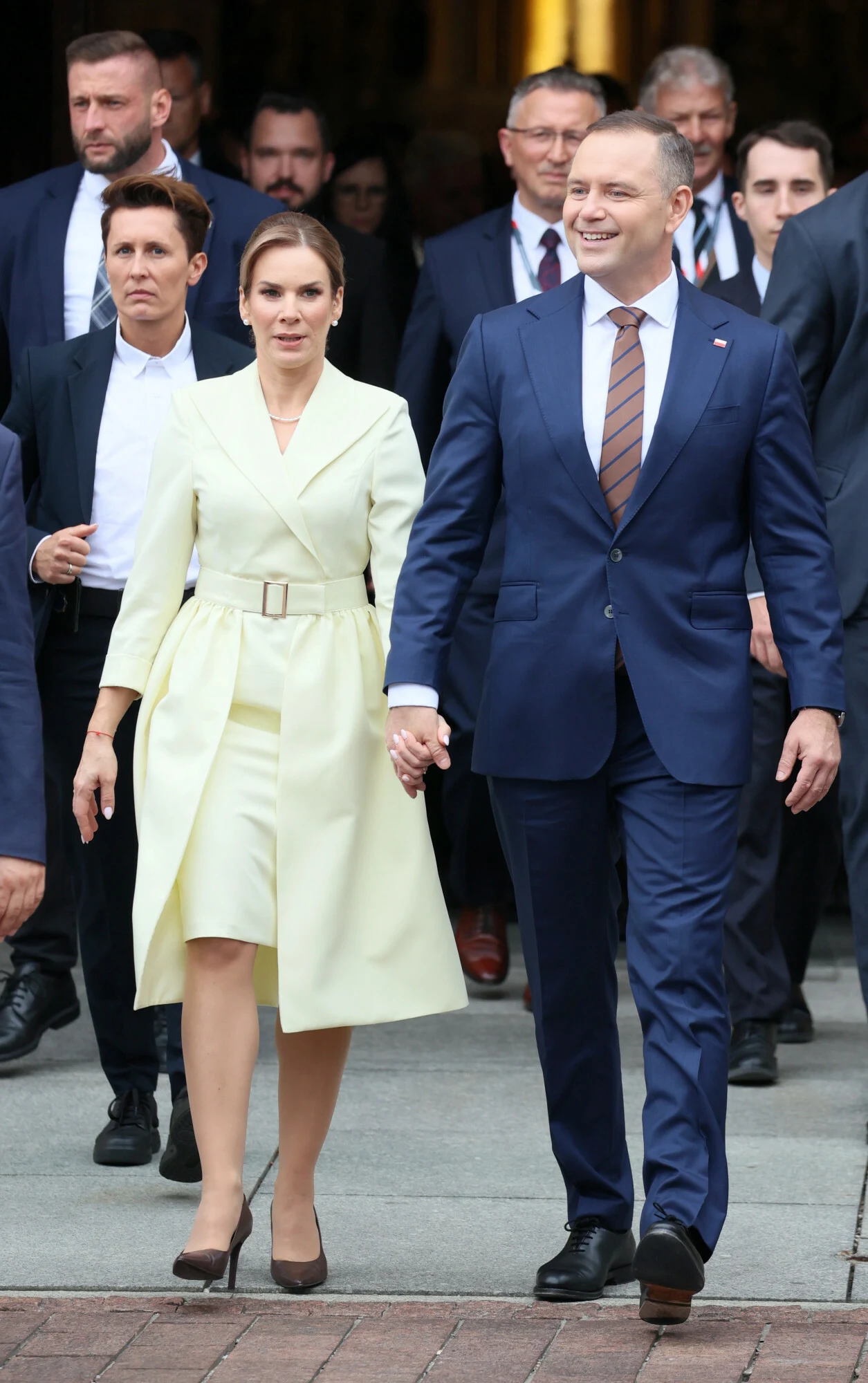 Para w eleganckich strojach, kobieta w jasnym płaszczu i mężczyzna w granatowym garniturze, trzymają się za ręce i idą w otoczeniu kilku osób.