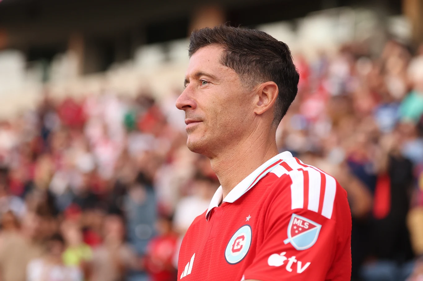 Robert Lewandowski Piłkarz w czerwonej koszulce z białymi paskami na ramionach i licznymi emblematami, stojący na stadionie podczas meczu, skupiony na wydarzeniach na boisku.