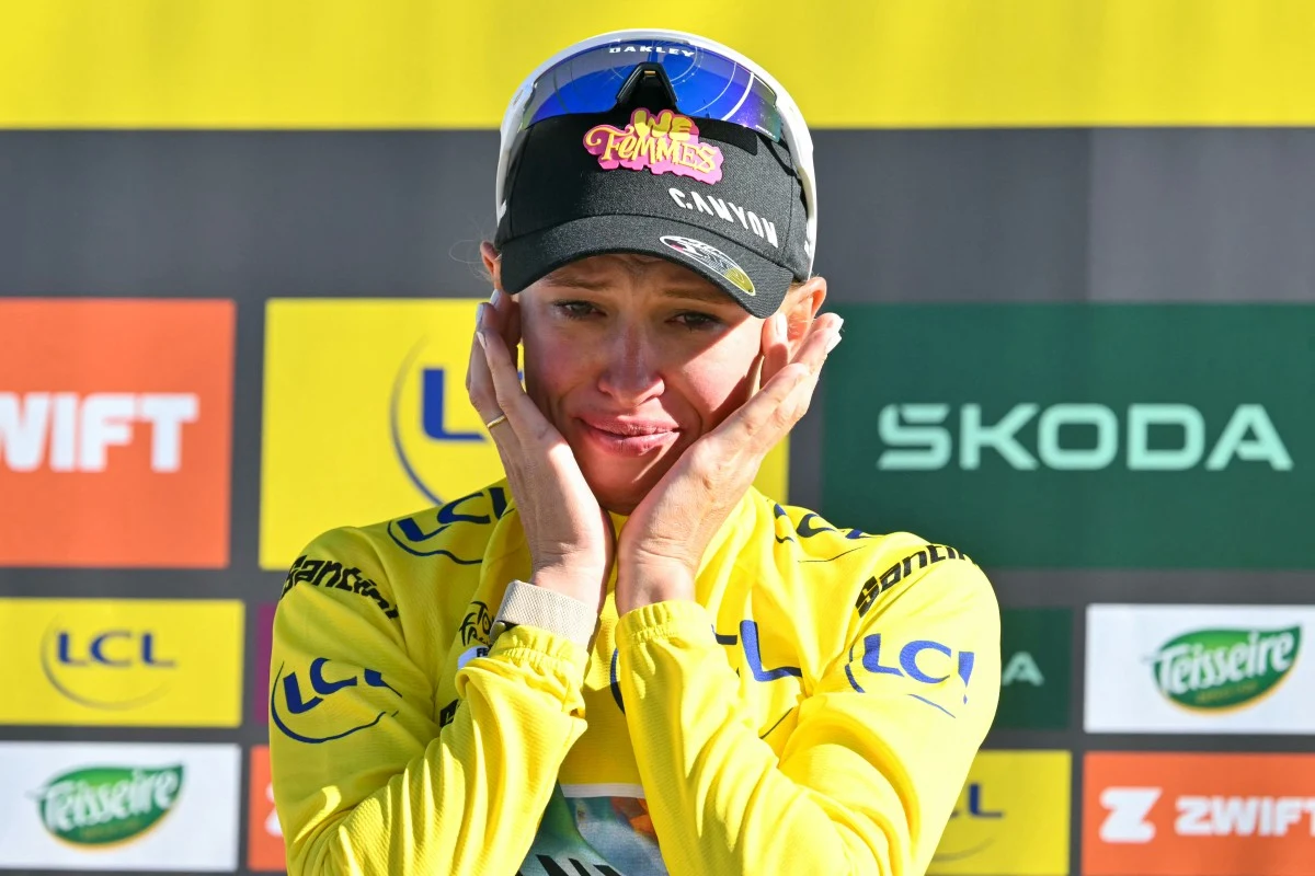 Katarzyna Niewiadoma-Phinney liderką Tour de France Kobieta w żółtej koszulce kolarskiej i czapce zespołowej trzyma dłońmi twarz, wyrażając silne emocje, na tle tablicy sponsorów wyścigu rowerowego.