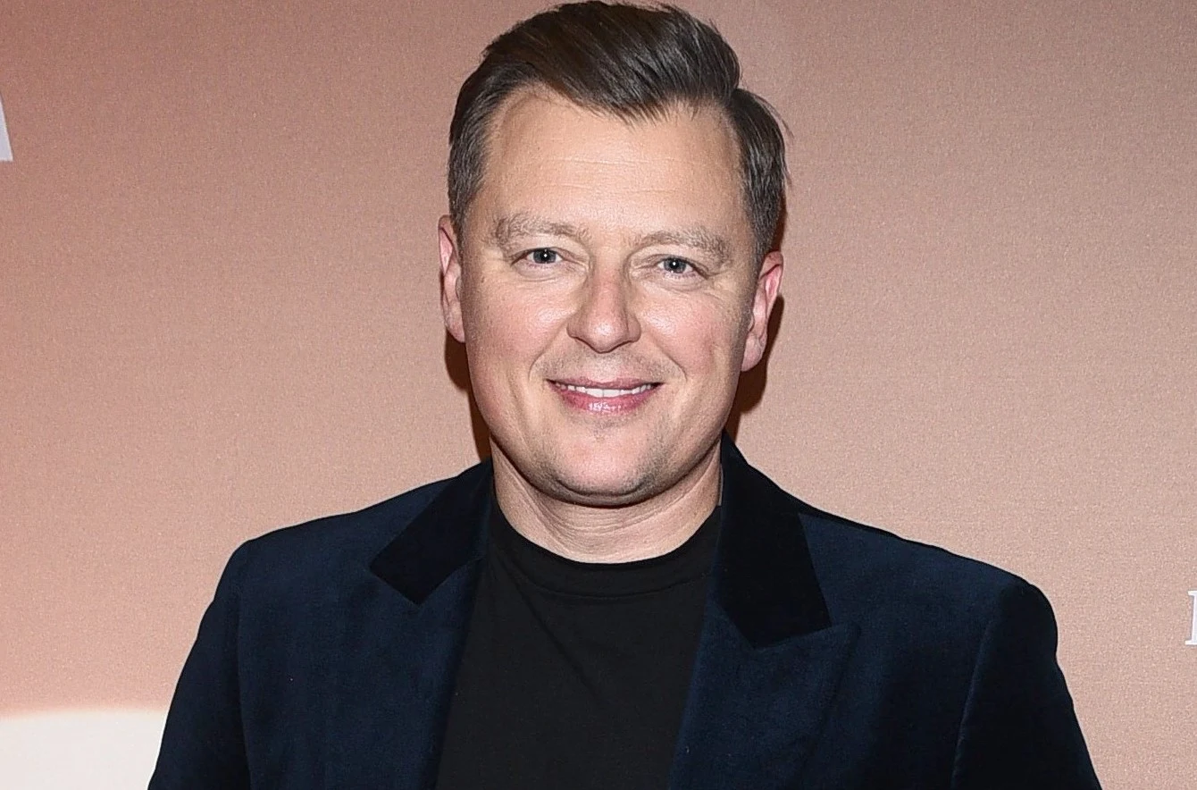Rafał Brzozowski Uśmiechnięty mężczyzna w granatowej marynarce i czarnym golfie na jednolitym tle.