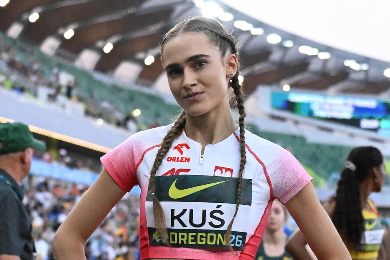 Anastazja Kuś, Eugene 2026 Lekkoatletka w biało-czerwonym stroju sportowym z numerem startowym, stojąca na stadionie lekkoatletycznym z rękami opartymi na biodrach, w tle widoczne trybuny i inni sportowcy.