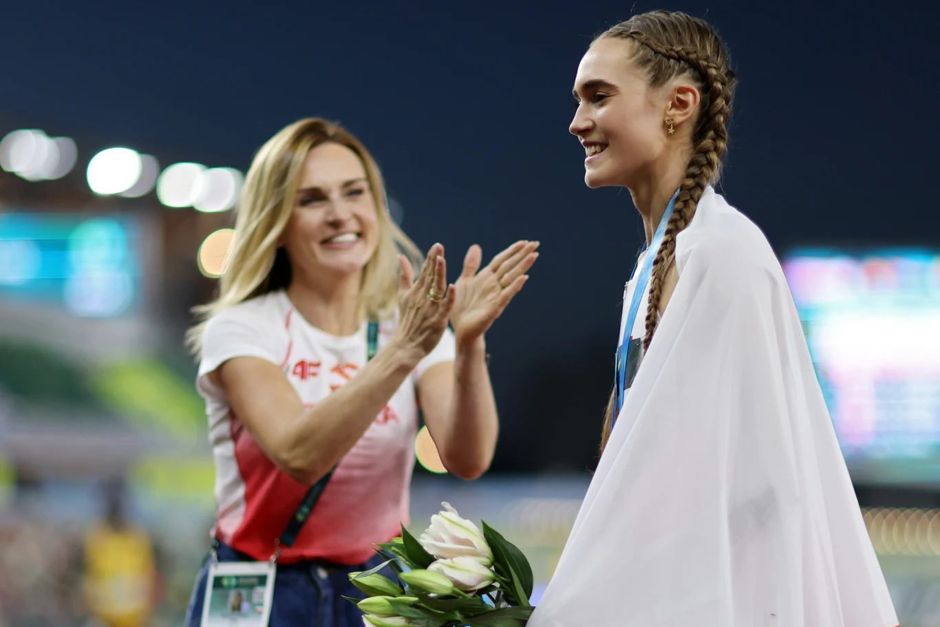 Z gratulacjami od razu po sukcesie ruszyła... mama Sportsmenka z medalem zawieszonym na szyi owinięta flagą otrzymuje gratulacje od uśmiechniętej kobiety i trzyma bukiet kwiatów.