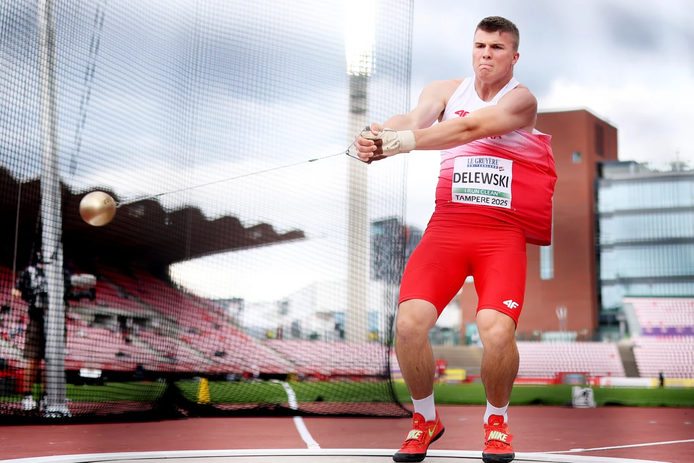 Lekkoatleta w czerwono-białym stroju wykonuje obrót podczas rzutu młotem na stadionie lekkoatletycznym, trzymając młot w obu dłoniach i skupiając się na wykonaniu rzutu.