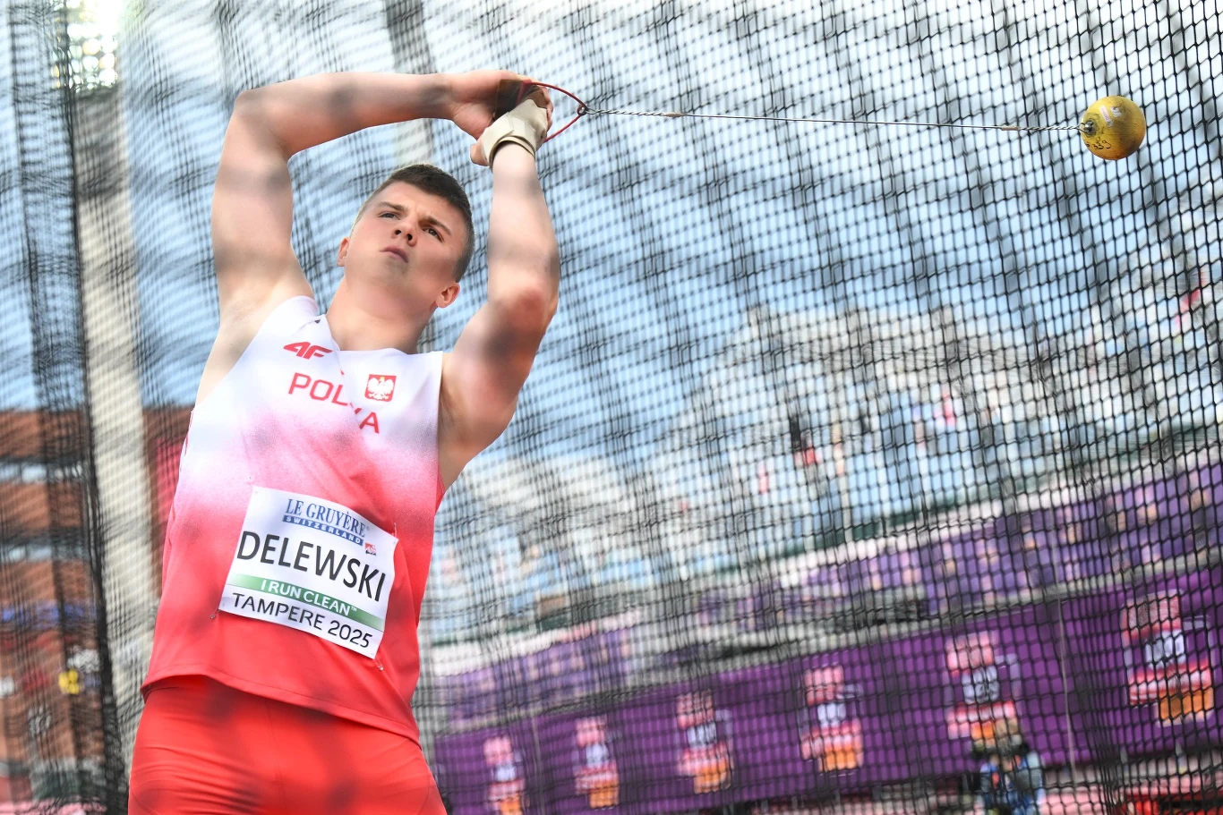 Lekkoatleta w stroju reprezentacji Polski wykonuje rzut młotem na stadionie lekkoatletycznym, w tle widoczne trybuny oraz siatka ochronna.