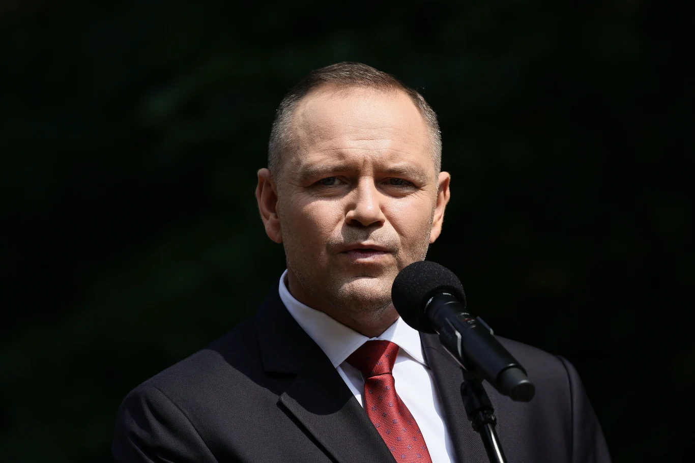 Mężczyzna w garniturze i czerwonym krawacie wygłaszający przemówienie do mikrofonu na tle ciemnej zieleni.