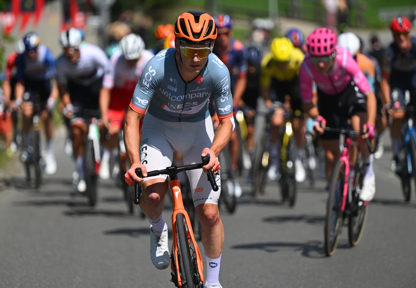 Magnus Sheffield Grupa kolarzy podczas wyścigu, jeden zawodnik w błękitnej koszulce i pomarańczowym kasku wyraźnie prowadzi peleton na asfaltowej trasie.