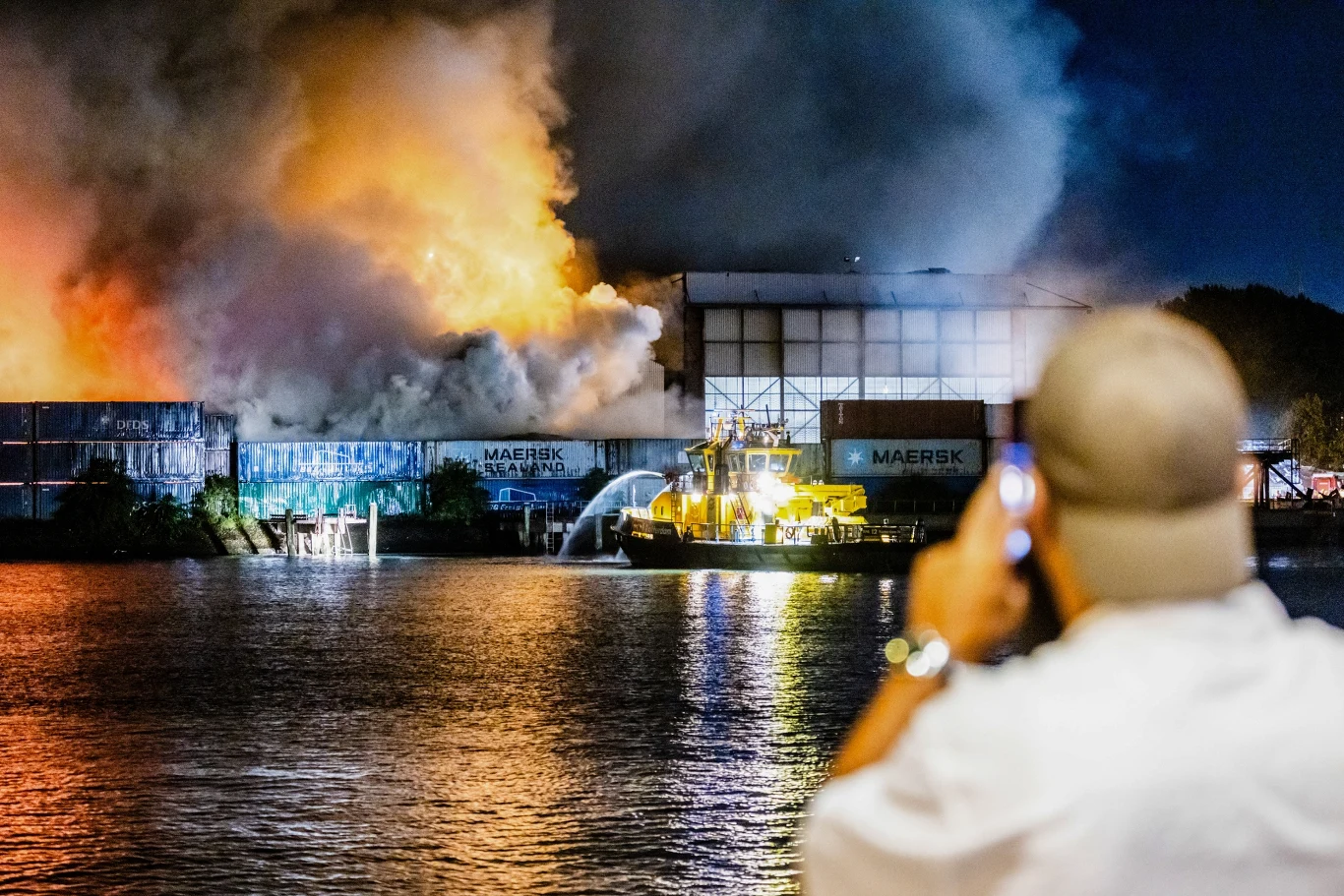 Duży pożar odpadów w Rotterdamie Płonący magazyn odpadów w Rotterdamie, dym, światło ognia, strażacy walczą z pożarem z łodzi, osoba dokumentuje.