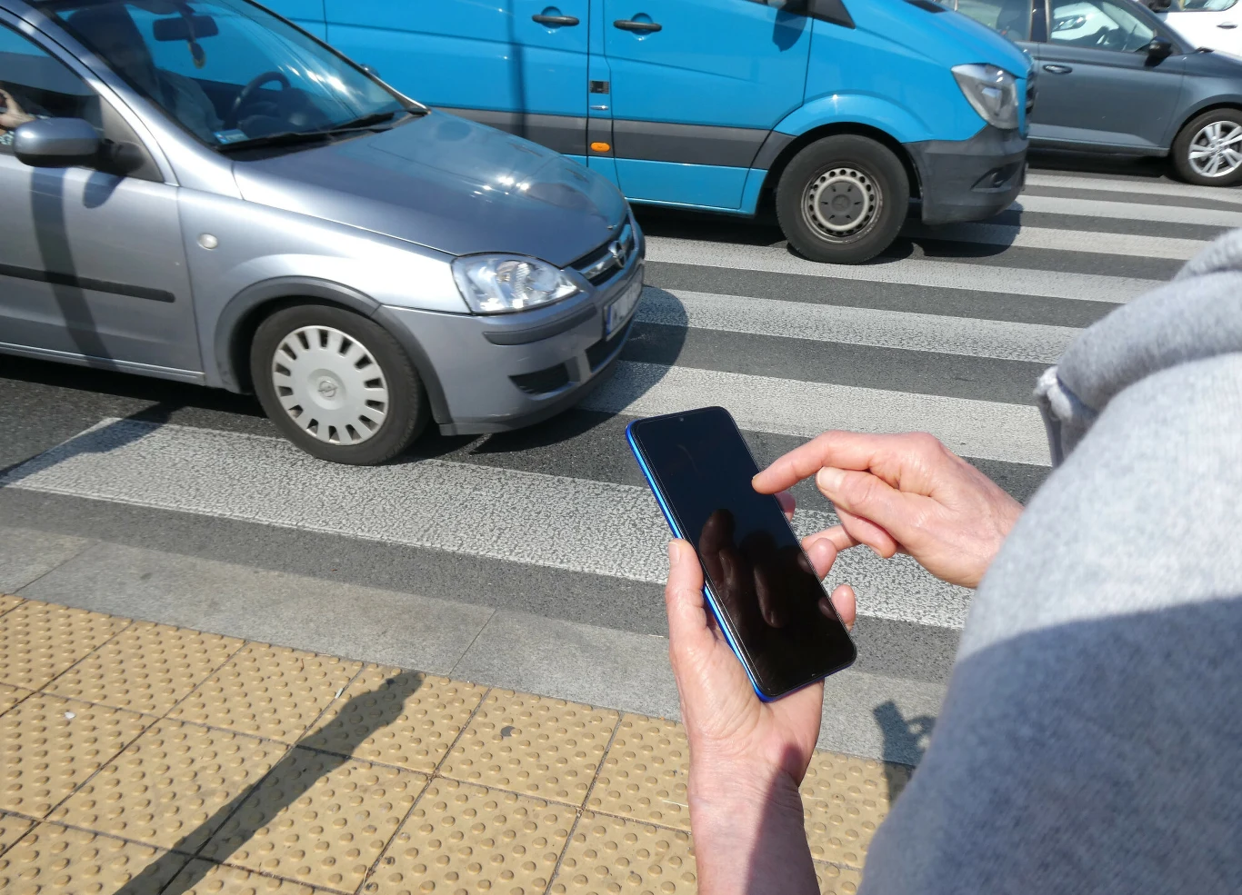 Szczególną ostrożność trzeba zachować np. w pobliżu przejść dla pieszych. Dotyczy to i kierowców i pieszych Osoba zbliżająca się do przejścia dla pieszych ze smartfonem, zatrzymane srebrny samochód i niebieski dostawczy na pasach.