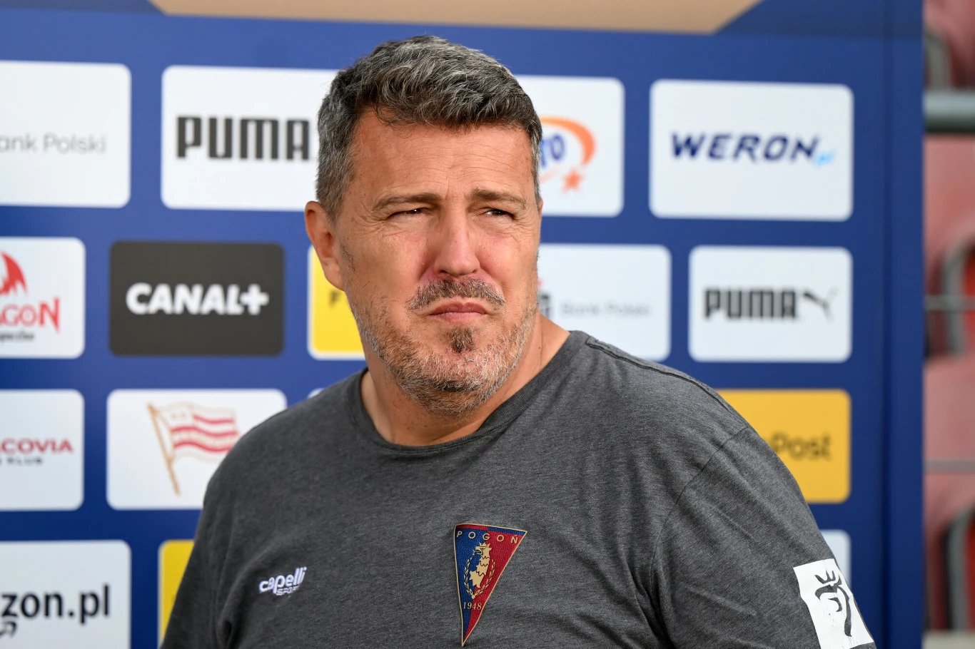 Mężczyzna w sportowej koszulce z herbem Pogoni Szczecin stoi na tle ścianki z logo sponsorów i partnerów klubu piłkarskiego.