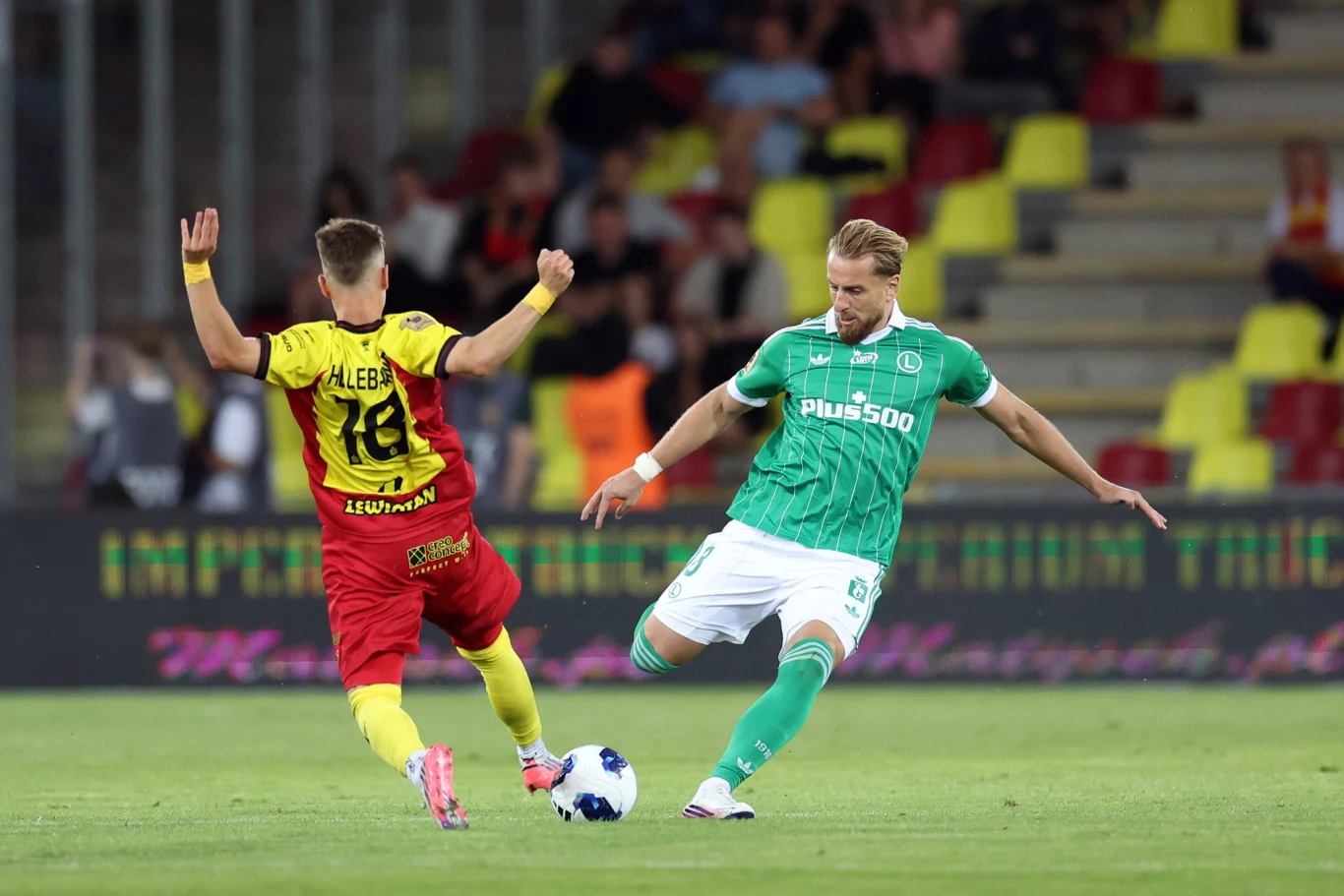 Korona Kielce - Legia Warszawa Dwóch piłkarzy w dynamicznej akcji podczas meczu, zawodnik w zielonym stroju próbuje odebrać piłkę zawodnikowi w żółto-czerwonym stroju, na tle pustych krzeseł stadionu.