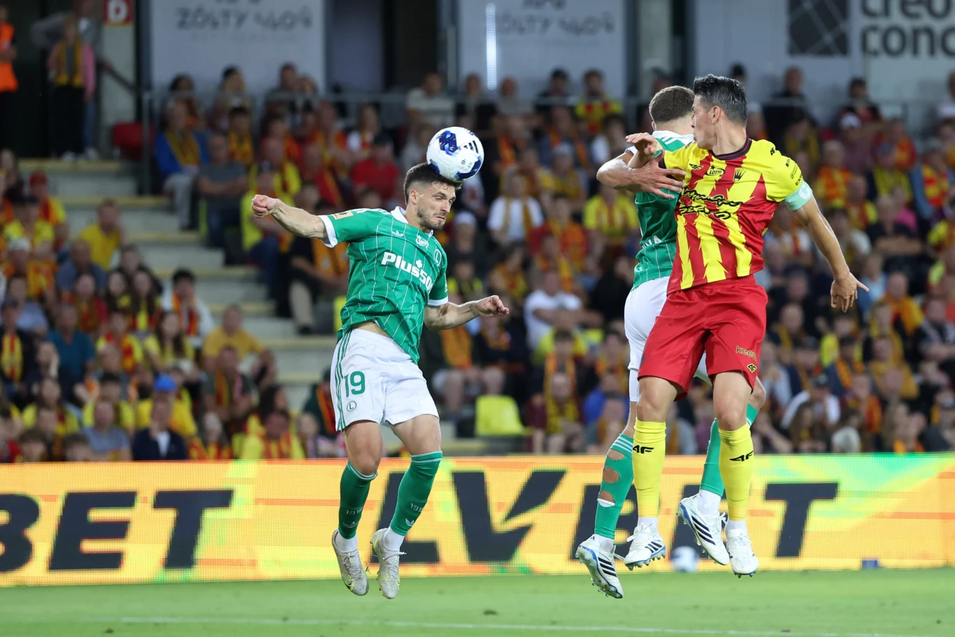 Korona Kielce - Legia Warszawa Dwóch piłkarzy rywalizuje o piłkę głową podczas meczu piłki nożnej, jeden w zielonej koszulce, drugi w żółto-czerwonym stroju, w tle widownia na stadionie.