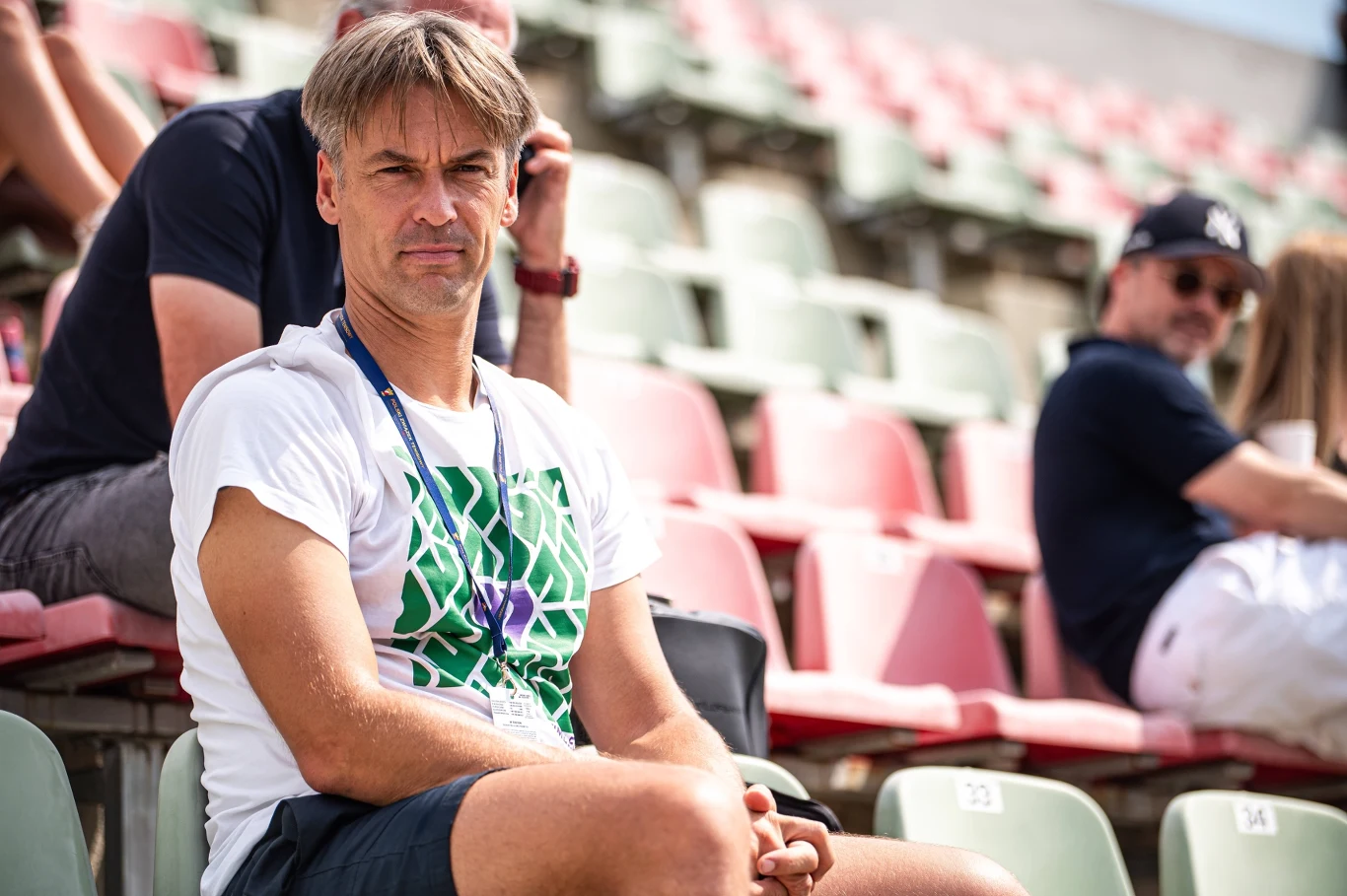 Mężczyzna w białej koszulce z zielonym wzorem siedzi samotnie na trybunach stadionu, patrząc przed siebie.
