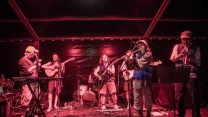 Zespół muzyczny wykonujący utwór na scenie, wokalista przy mikrofonie, gitarzyści, perkusista oraz dwóch muzyków grających na trąbkach w intensywnym, czerwonym oświetleniu scenicznym.