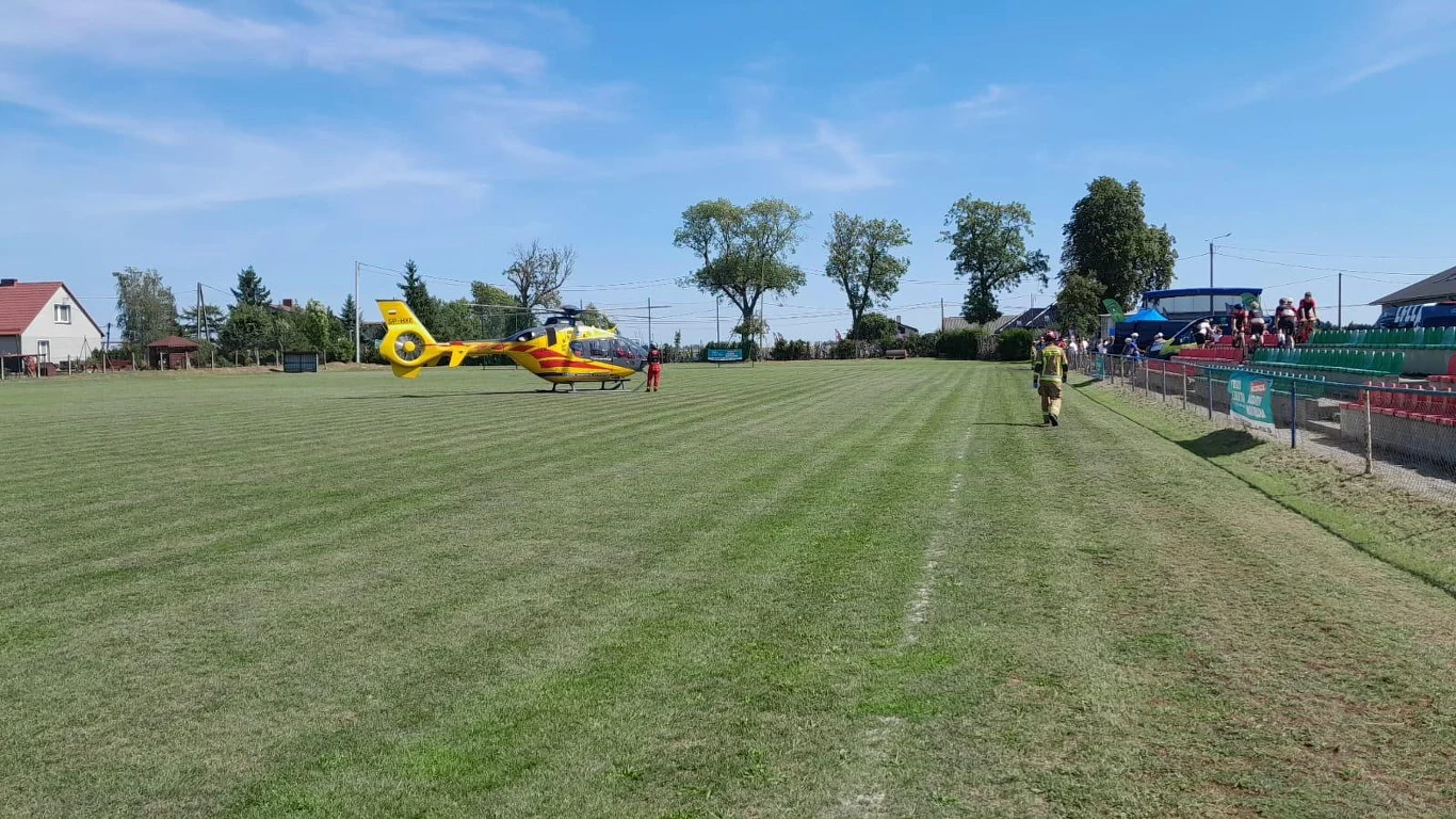 Żółty helikopter ratunkowy ląduje na środku boiska sportowego, w pobliżu trybun z grupą ludzi oraz samotnie idącym strażakiem w stronę maszyny.