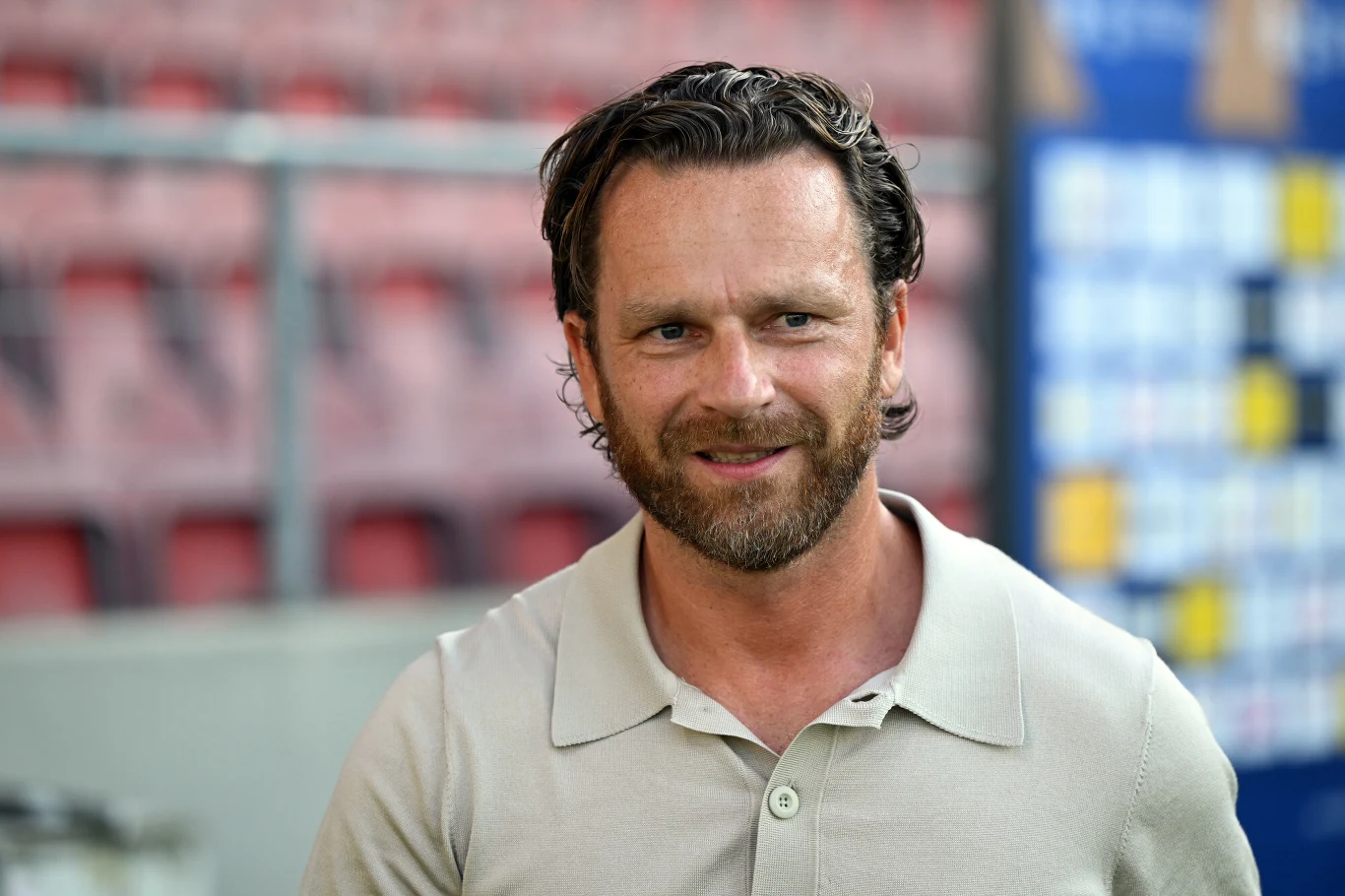 Bartosz Grzelak Mężczyzna z krótkimi, ciemnymi włosami i brodą, ubrany w jasną koszulkę polo, patrzy z lekkim uśmiechem w stronę obiektywu; za nim rozmyte trybuny i tablica z kolorowymi prostokątami.