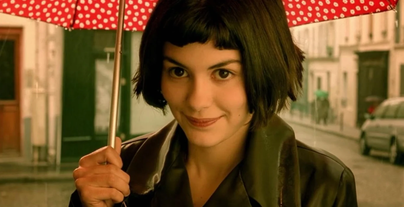 Audrey Tautou jako Amelia Młoda kobieta o krótkich, ciemnych włosach uśmiecha się lekko, trzymając czerwony parasol w białe kropki na tle miejskiej ulicy.