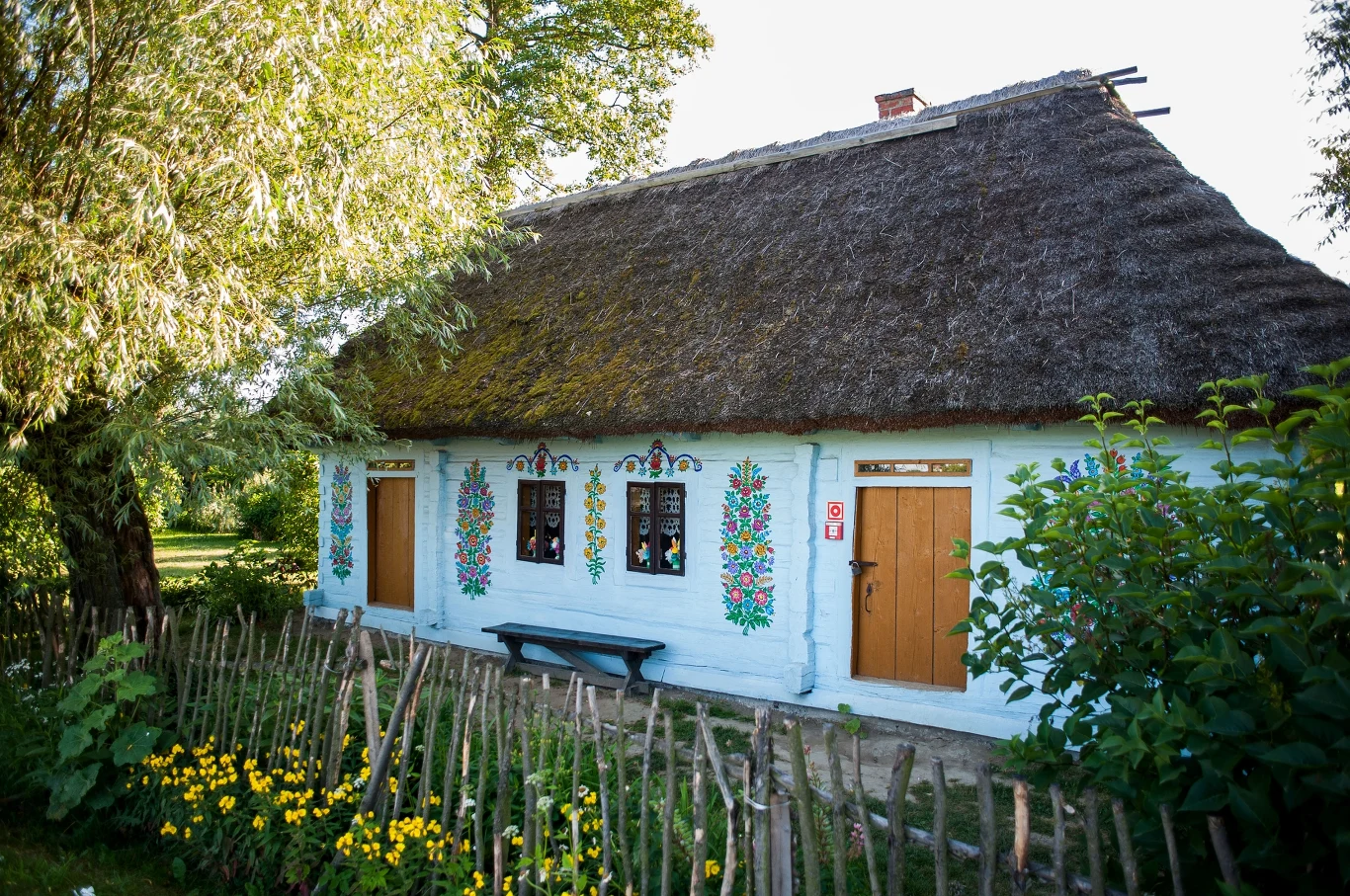 Mały wiejski domek o bielonych ścianach, zdobionych kolorowymi malowidłami ludowymi, z dachem pokrytym strzechą, otoczony drewnianym płotem i kwitnącymi kwiatami.