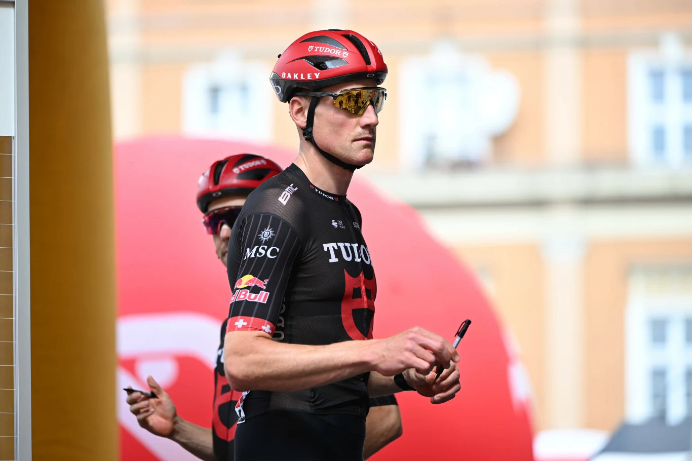 Stefan Kung Kolarz w czarnym stroju zespołu Trek-Segafredo z czerwonym kaskiem i okularami stoi w wyprostowanej pozycji, trzymając marker w dłoni; za nim widoczny drugi zawodnik tej samej drużyny na tle dużego czerwonego balonu.