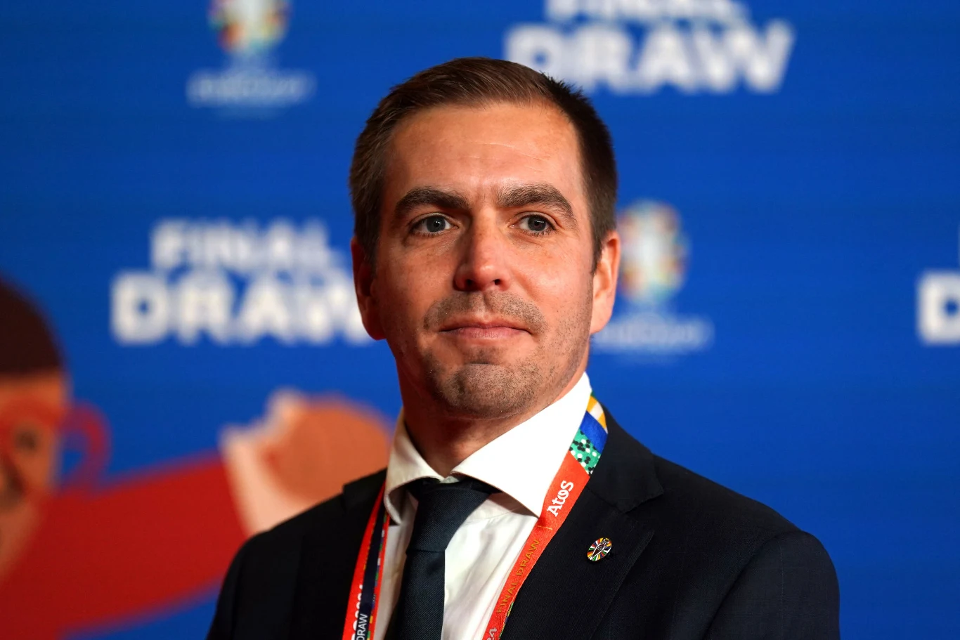 Philipp Lahm Mężczyzna w garniturze z identyfikatorem na szyi stoi na tle niebieskiej ścianki z kolorowym nadrukiem i napisem 'FINAL DRAW'.