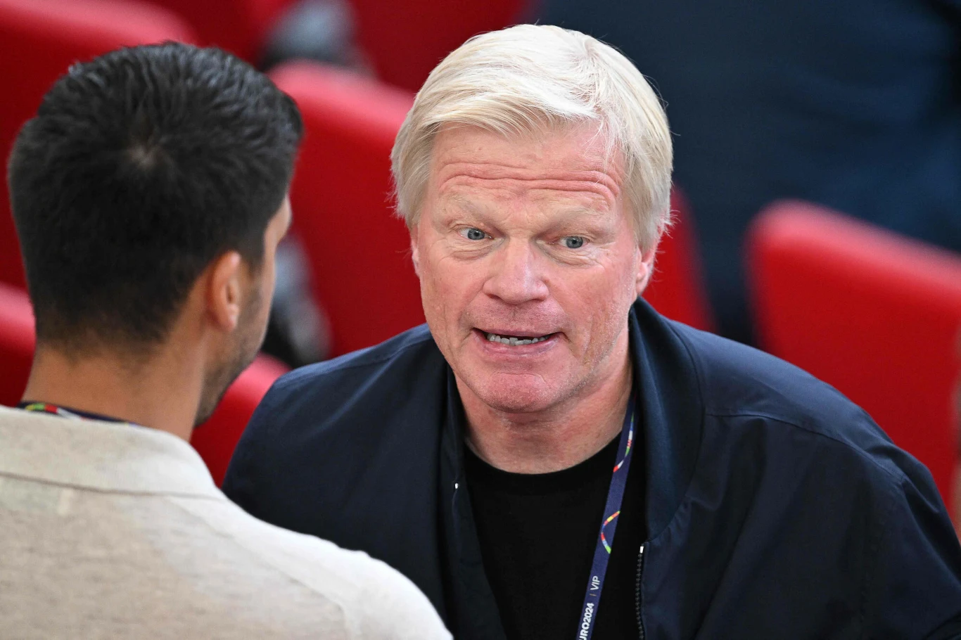 Oliver Kahn Dwóch mężczyzn rozmawia twarzą w twarz na tle czerwonych siedzeń, jeden z nich ma jasne blond włosy i ubrany jest w ciemną kurtkę.