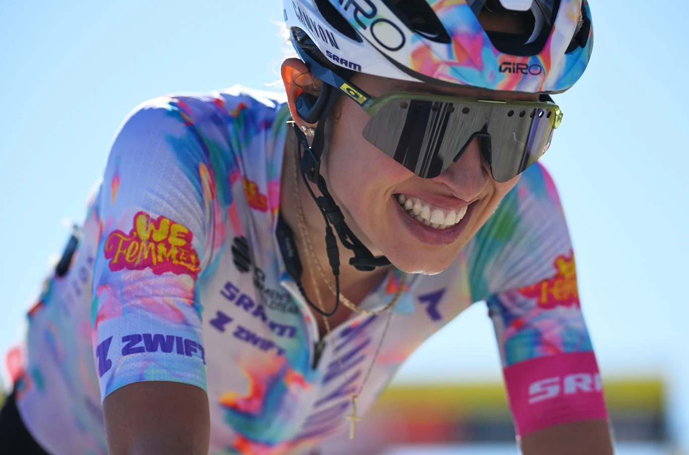 Katarzyna Niewiadoma-Phinney Uśmiechnięta kolarka w sportowej koszulce w kolorowe wzory i okularach przeciwsłonecznych podczas wyścigu rowerowego.